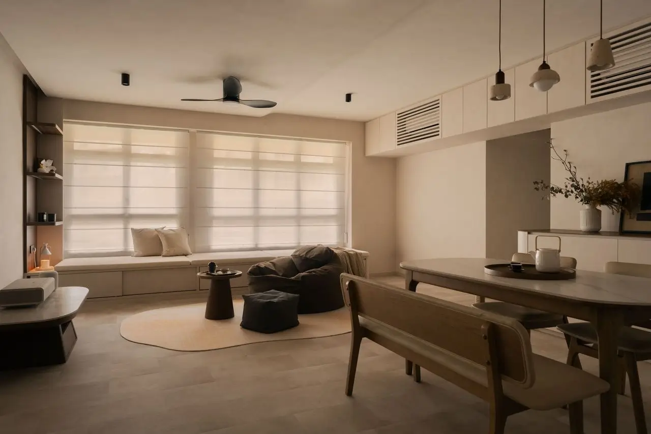 Warm, minimalist open-plan living and dining interior with a neutral beige palette, large windows dressed with Roman shades, built-in window seating, soft textured furnishings, bean bag lounge chairs, a light area rug, and a curved wooden coffee table. The space features custom cabinetry, concealed storage, a streamlined dining area with solid wood furniture, modern pendant lighting, a ceiling fan, and subtle natural finishes that create a calm, contemporary aesthetic.
