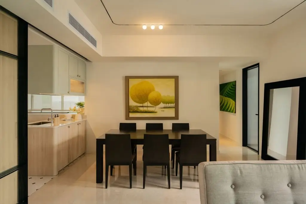 Modern contemporary condo interior design in Singapore featuring an open-plan dining and kitchen area with minimalist cabinetry, a six-seater dining table, neutral colour palette, framed artwork, and warm ambient lighting that creates a bright and elegant living space.